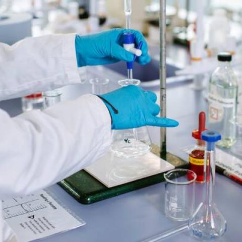 Person in white coat and blue gloves putting liquid in a glass beaker in a lab