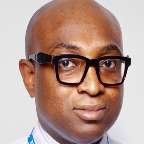 Headshot of Ade Williams MBE looking to camera wearing glasses, a shirt and a NHS lanyard