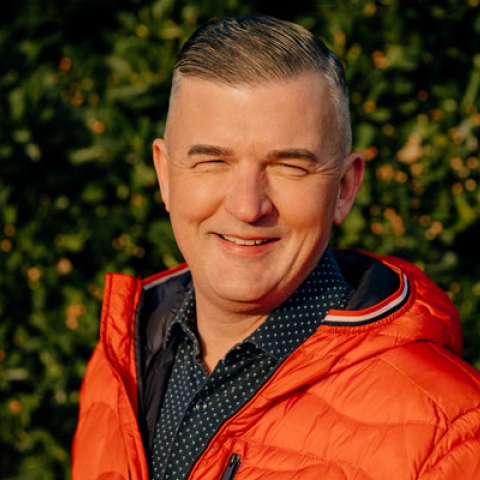 Headshot of Craig Jones MBE wearing orange jacket and smiling to camera with greenery backdrop