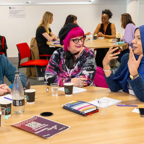 A group of women seated around a table engaged in a lively discussion at a professional event. One woman in a blue hijab is speaking animatedly, using hand gestures, while two others, one wearing glasses and a blue sweater and the other with vibrant pink hair and a patterned outfit, listen attentively with smiles. The table is filled with notebooks, coffee cups, and event materials. In the background, other attendees are seen networking and conversing.