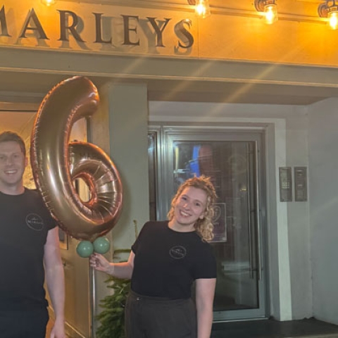 Two University of Portsmouth graduates standing in front of their restaurant, Marley's with a big '6' balloon smiling to camera