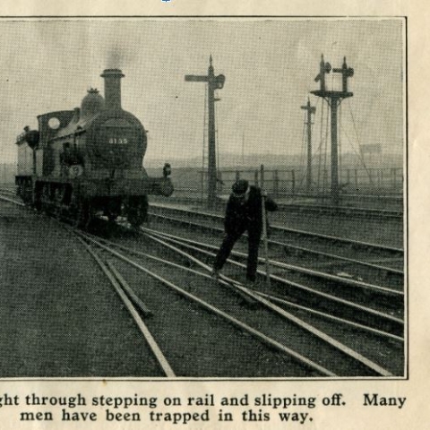 Historic black and white photo of a steam rail engine in background on the tracks and a 1930s railway worker on the track posing as if his foot is caught in a track