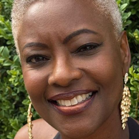 Image of Samantha Tross smiling to camera wearing gold dangly earrings against greenery backdrop