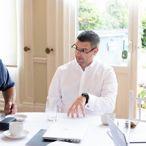 Two men at a table with their laptops talking