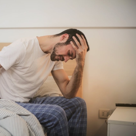 Man sitting on bed with one hand on his head and one on his head, looking in pain