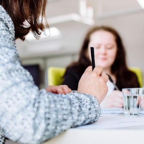 a close up image of a hand holding a pen, with an out of focus person in the background