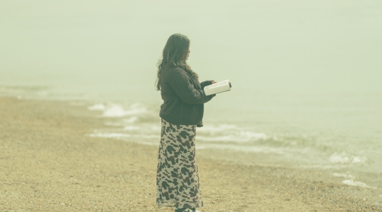 photograph of woman reading book on the beach whilst facing the sea