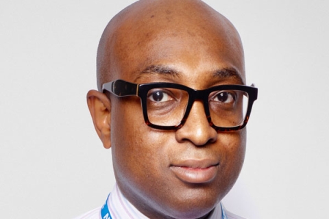 Headshot of Ade Williams MBE looking to camera wearing glasses, a shirt and a NHS lanyard