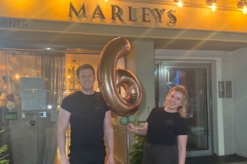 Two University of Portsmouth graduates standing in front of their restaurant, Marley's with a big '6' balloon smiling to camera