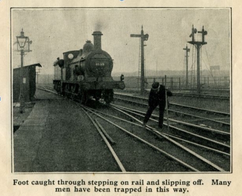 Historic black and white photo of a steam rail engine in background on the tracks and a 1930s railway worker on the track posing as if his foot is caught in a track