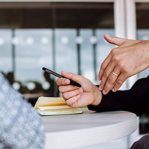 Two people discussing notes, one holding a pen and pointing at the notebook on a white table