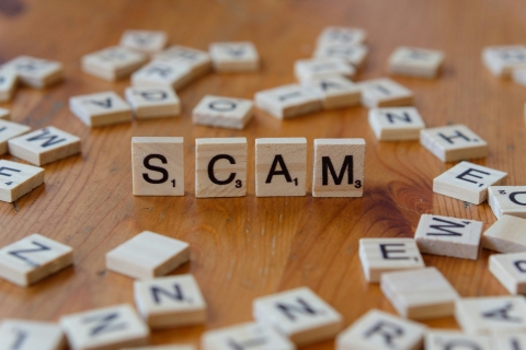 An array of Scrabble letter tiles on a wooden table top with four tiles standing upright to spell the word SCAM.  Photo credit Unsplash