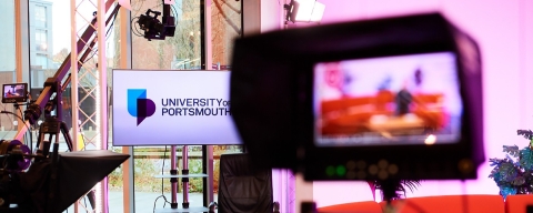 A picture of podcasting hardware in a recording studio with a screen in the background with the university of portsmouth logo displayed