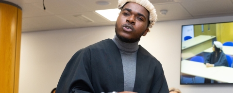 A man standing in a replica law court wearing a solicitors robe a white periwig