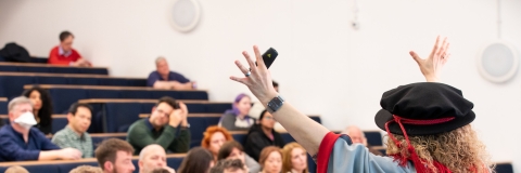 Professor Banu in academic robes with her arms up, delivering a lecture in front of an audience.