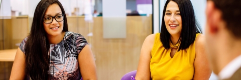 Two women in a meeting, sat across a table from a man with his back to the camera