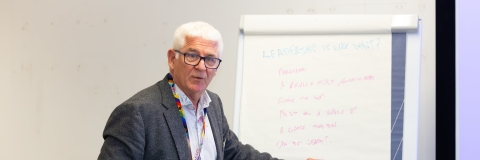 Male with white hair with his hand pointing to a flip chart in a lecture room