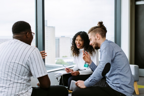 Three students sat laughing and smiling holding notebooks and pens