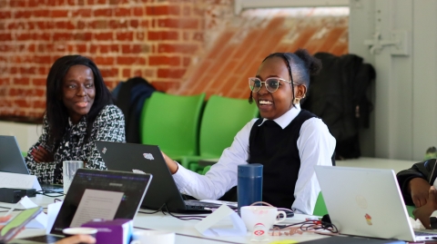 A student sat at a table wearing a black and white top talking to other students 