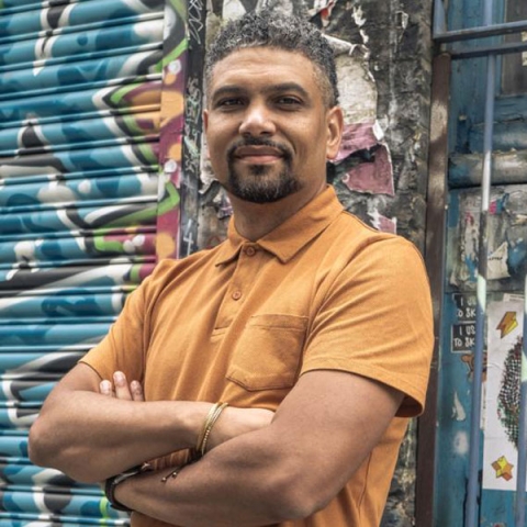 Profile image of alumnus Leon Mann smiling to camera wearing orange polo against graffiti wall