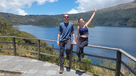 Two individuals smile for a photo on a bridge with a scenic view of a lake in the background.