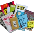 a collection of zines stacked on one another including one called obsession and one with a dog