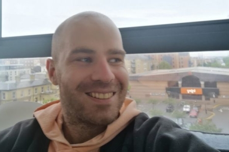 Male with beard and shaved head wearing an orange hoodie with his back against a window 