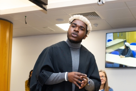 A man standing in a replica law court wearing a solicitors robe a white periwig