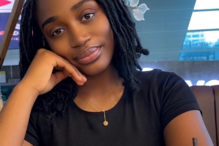 Female student sitting at a table. Wearing black tshirt and shoulder length hair. Behind her is a lamp and in front on the table is a bag, cup and glasses.