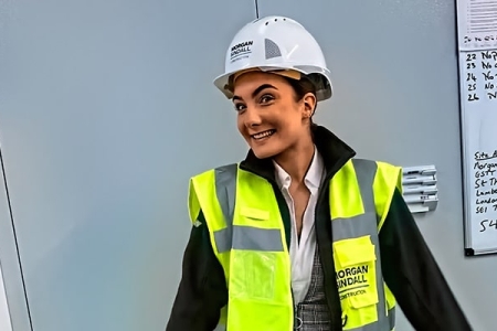 A woman in viz jacket and hard hat, standing in front of a whiteboard with writing on it, smiling at the camera