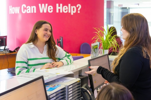 How to help is written on the wall in the background. In the forefront is a student standing at a student support counter seeking help from a member of staff
